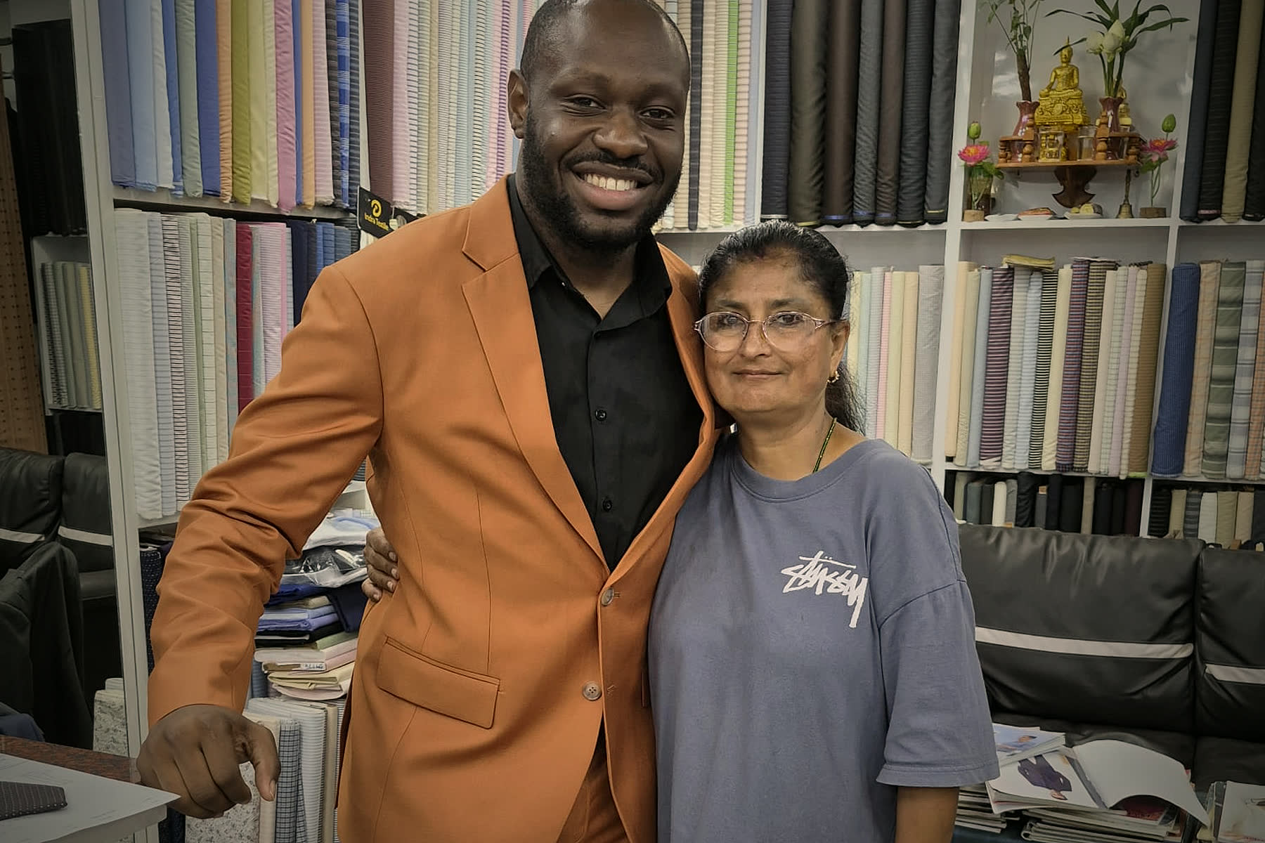 A guest in a freshly made orange suit, next to one of the tailors in the shop